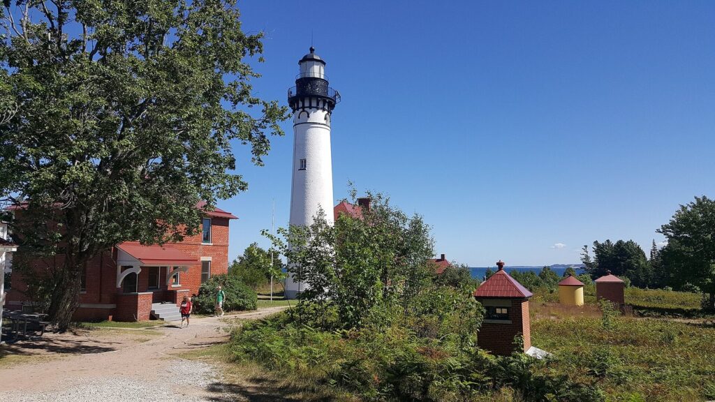 White Au Sable lighthouse tower beside red-brick keepers' buildings under a blue sky