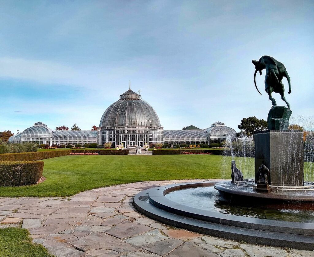 Domed glass conservatory beyond a lawn and a fountain with a bronze statue under a blue sky