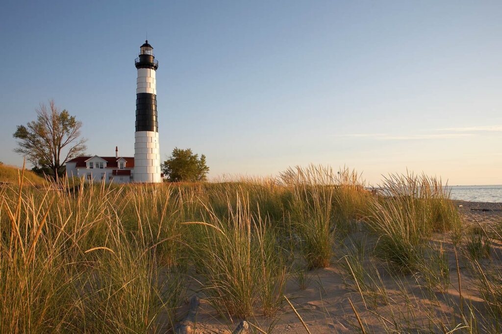 Black-and-white Big Sable Point Lighthouse and its red-roofed keeper's house behind dune grass in low evening light