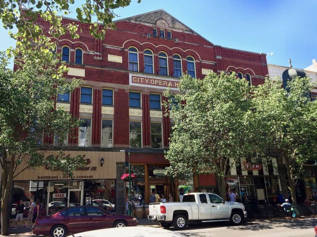 Red-brick City Opera House facade on Front Street with arched windows, trees and parked cars