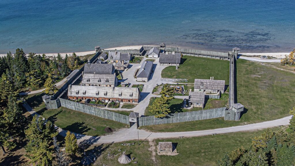 Aerial view of a wooden stockade fort with log buildings on a grassy point beside blue water