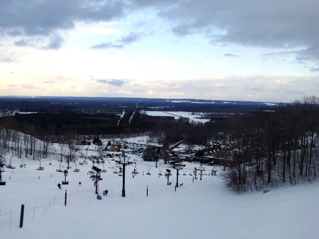 View down a groomed ski slope to chairlifts and a resort village surrounded by snowy forest