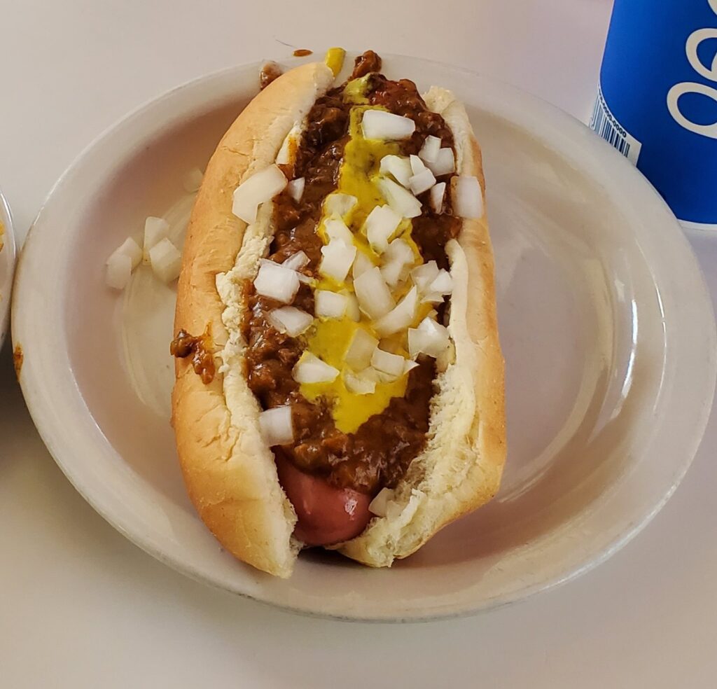 Coney dog topped with beanless chili, mustard and chopped onions on a white plate