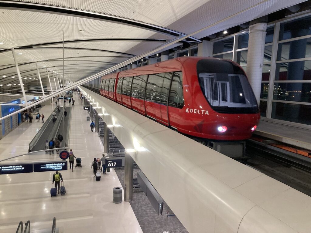 Long airport concourse with travelers, moving walkways and a red Delta tram running overhead at Detroit Metro
