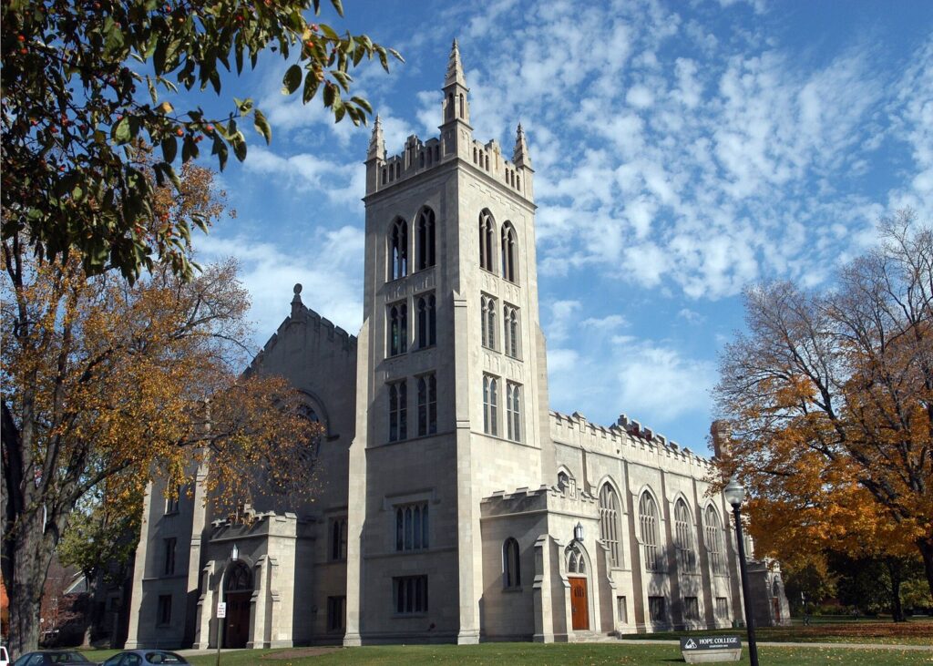 Gothic-style stone chapel with a tall square tower under a blue sky with autumn trees