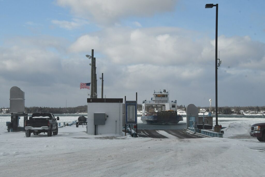A car ferry approaching a snowy dock beside a ticket booth and parked pickup truck in winter