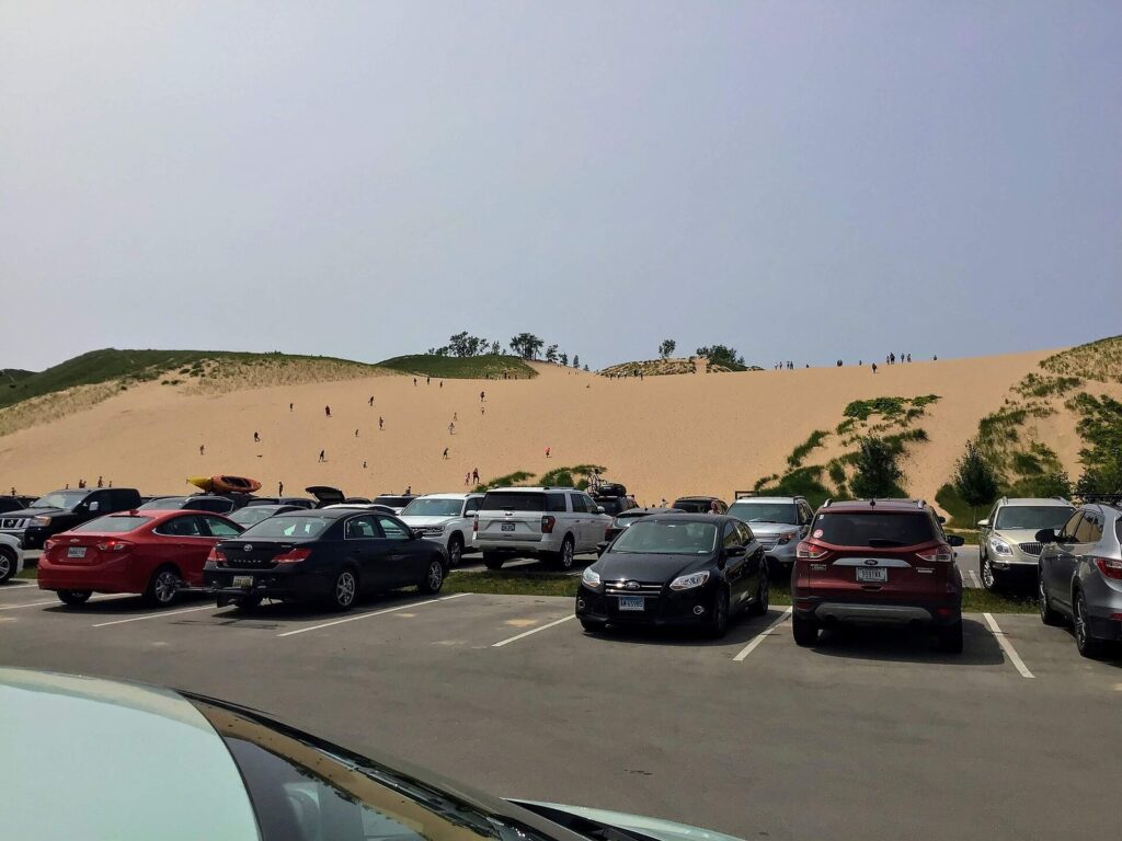 Parking lot full of cars in front of the Dune Climb, a large sand slope with tiny figures climbing it