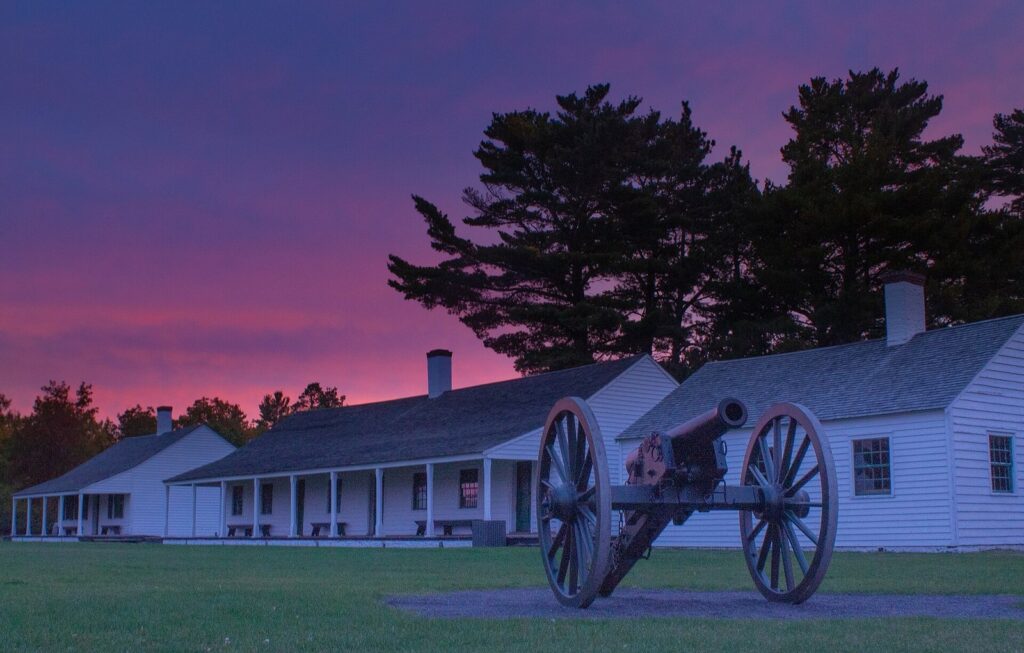 White wooden fort buildings and a cannon on a lawn beneath dark pines against a pink dawn sky