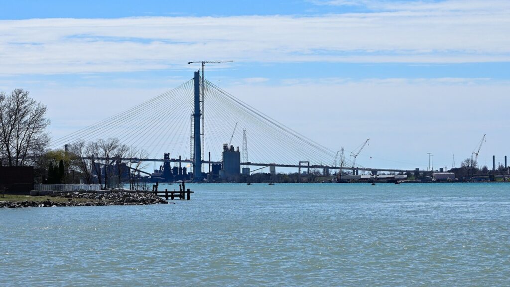 Tall white cable-stayed bridge tower rising over the Detroit River with industrial buildings on the far shore