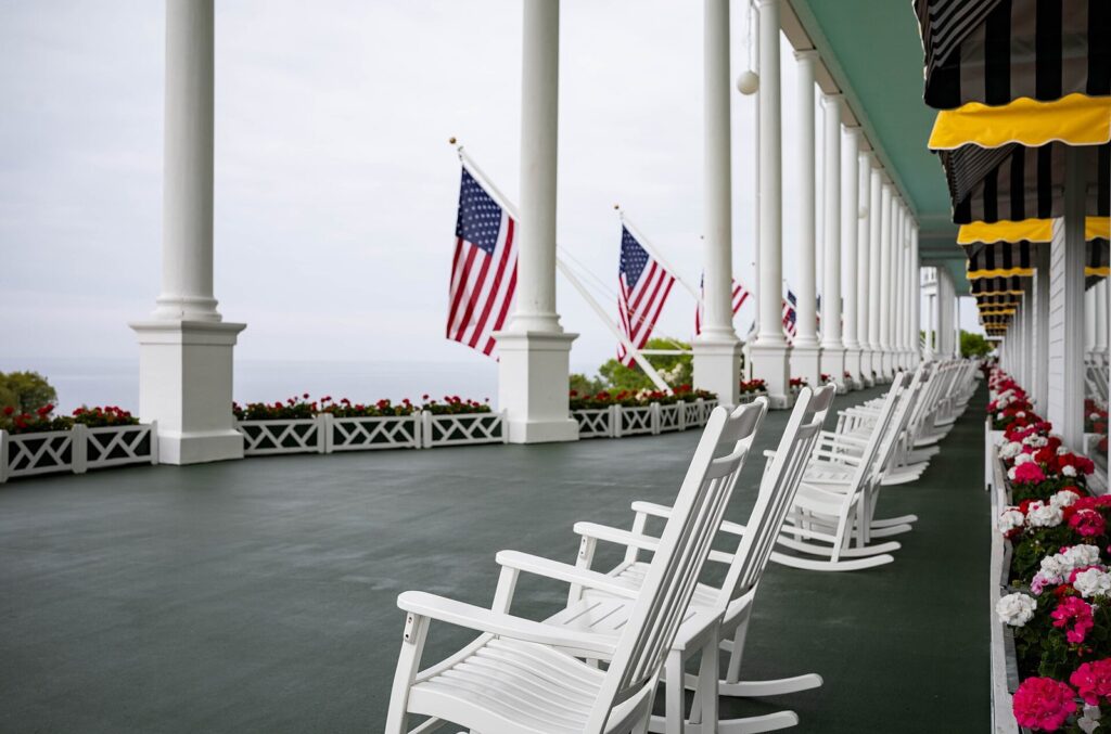 White rocking chairs lined along a long covered porch with flags and red flower boxes overlooking the water