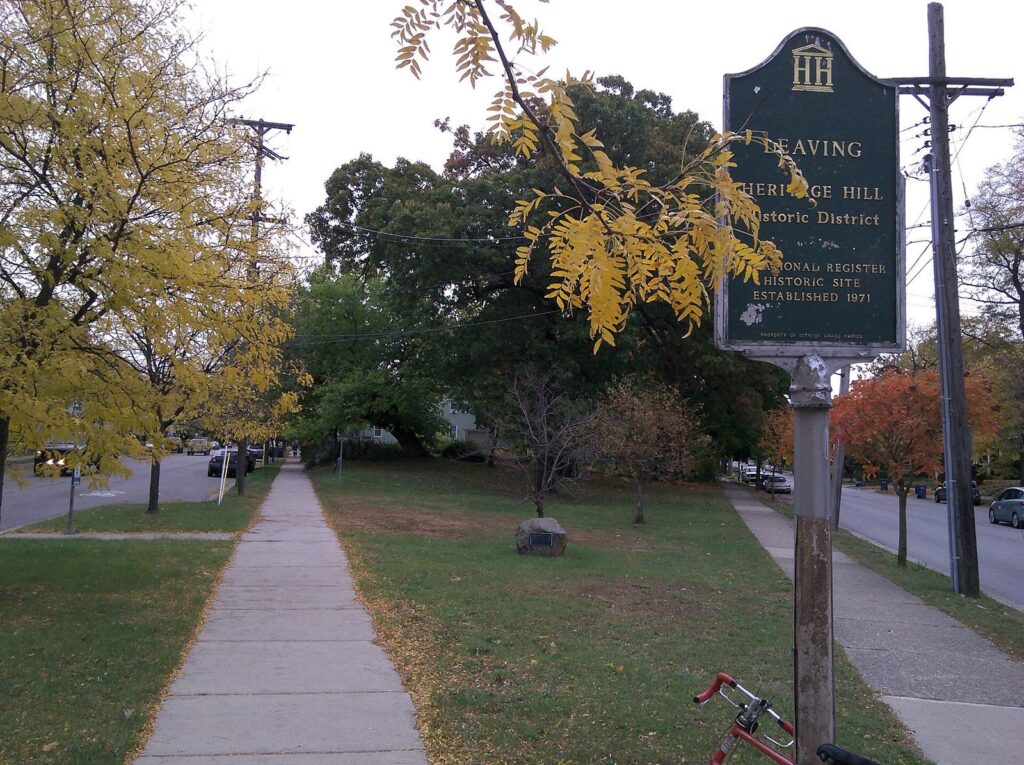 Tree-lined sidewalk in fall beside a green historic-district sign in the Heritage Hill neighborhood