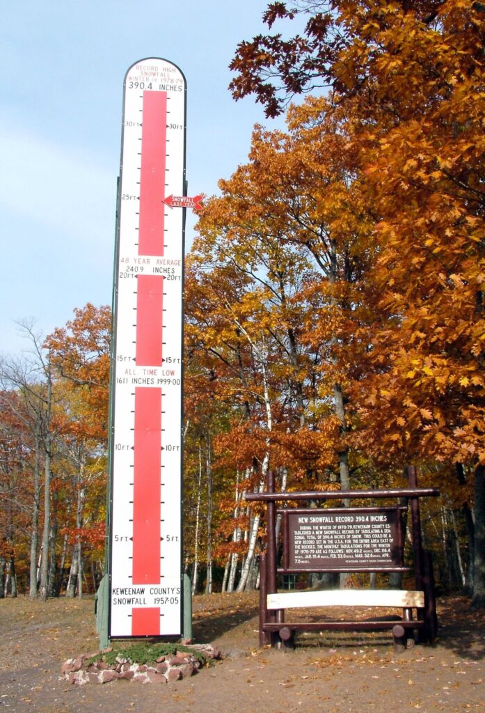 Tall red-and-white snow gauge sign marking record and average snowfall in feet, beside autumn trees