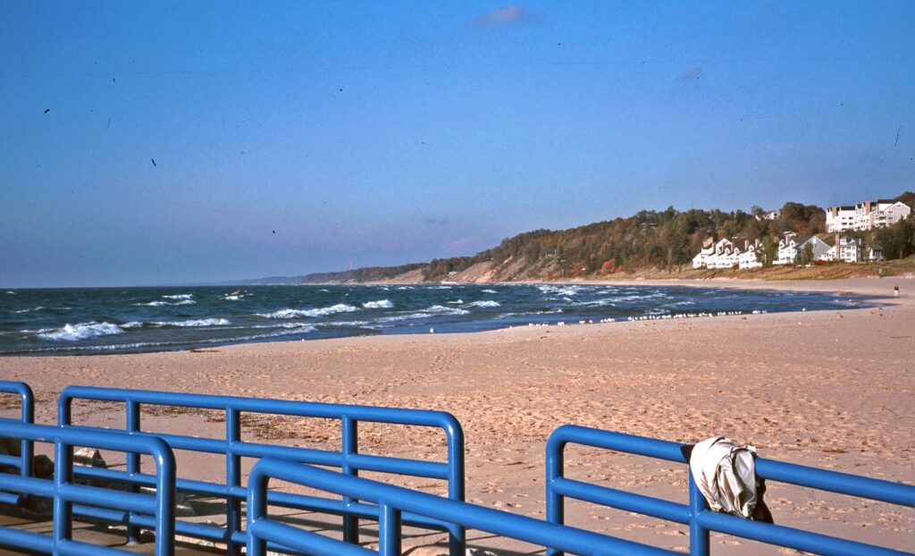 Wide sandy beach on Lake Michigan with blue railings in the foreground and houses on a wooded dune behind