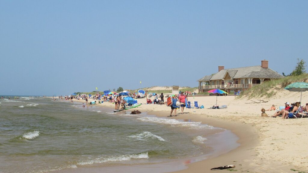 Busy summer beach with umbrellas and swimmers in the surf, with a historic beach house on the dunes behind