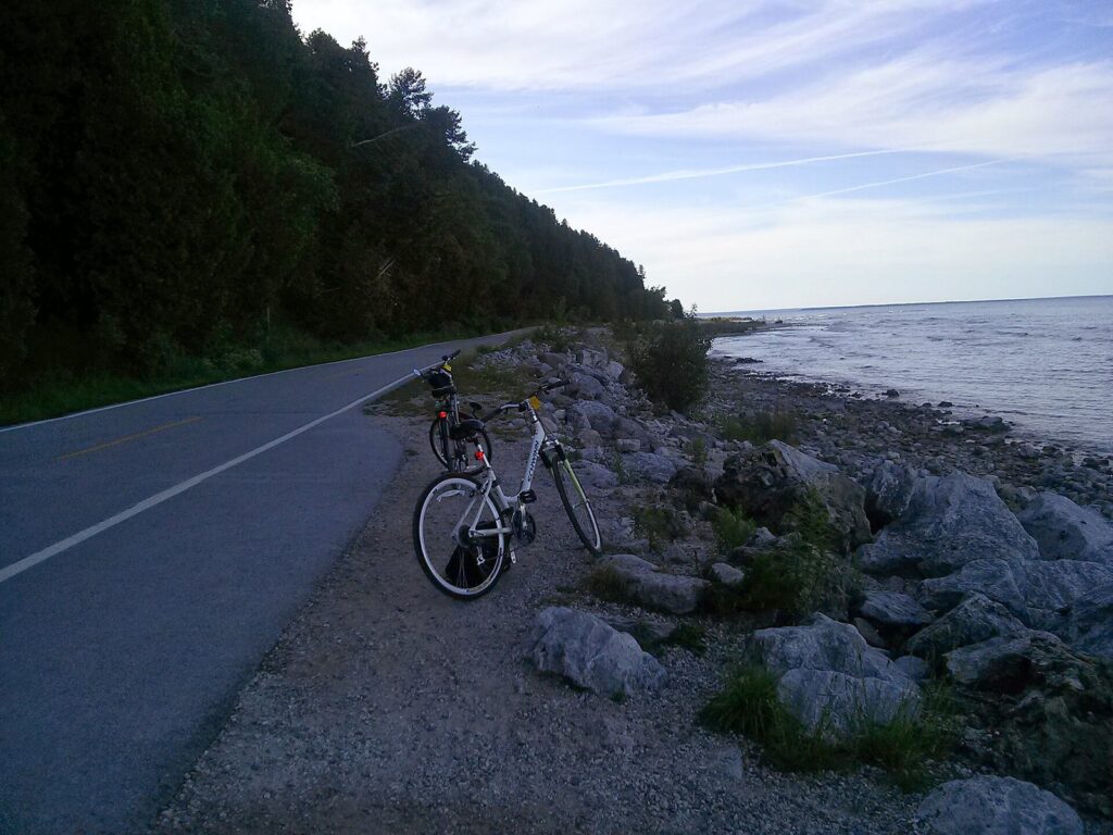 Two rental bikes parked beside the lakeshore road with a wooded bluff on the left and open water on the right
