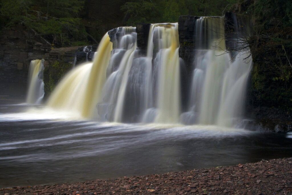 Manabezho Falls, a wide curtain of tea-colored water pouring over a rock ledge into a pool