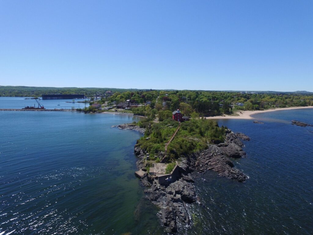 Aerial view of a red lighthouse on a rocky wooded point surrounded by blue Lake Superior water