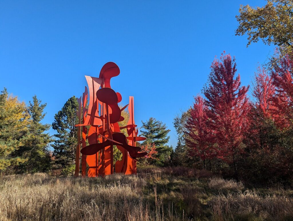 Red abstract steel sculpture in a meadow ringed by fall-colored trees under a blue sky at Meijer Gardens