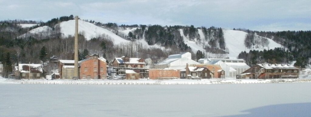 Snowy ski runs on a hillside above a frozen canal and brick industrial buildings with a tall chimney