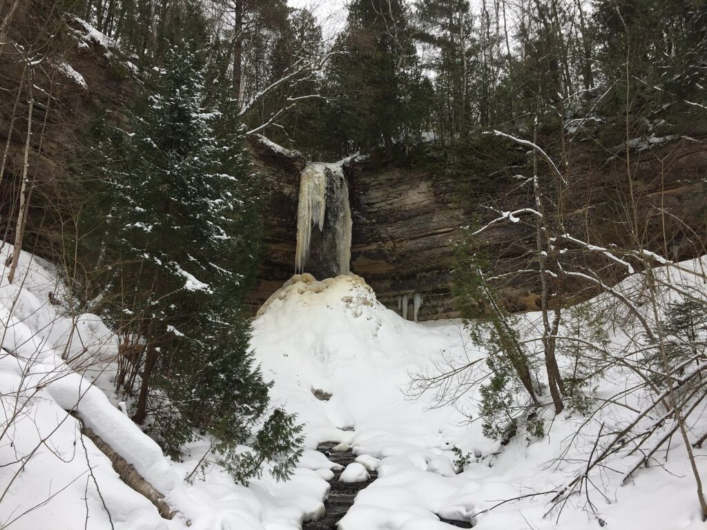 Munising Falls frozen into a column of ice above a snow mound, surrounded by snowy evergreens