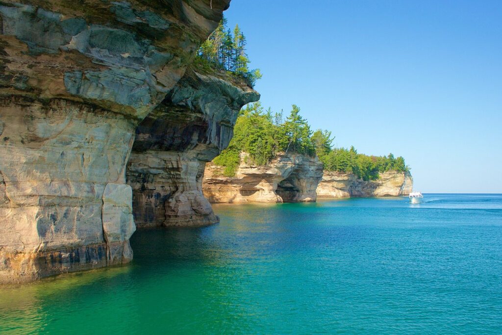 Tall sandstone cliffs and overhangs above turquoise water, with a tour boat far down the shoreline