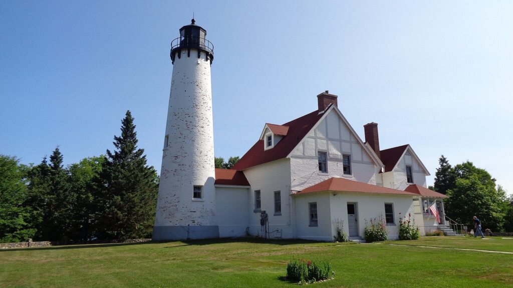 White brick lighthouse tower attached to a red-roofed keeper's house on a green lawn under a blue sky