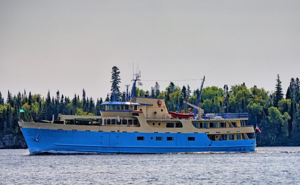 Blue-and-white passenger ship Ranger III cruising past a spruce-covered shore on calm water