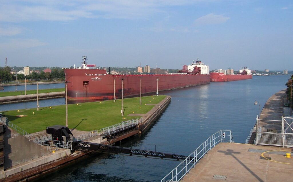 Long red freighter in a lock chamber beside a grassy wall, with more ships on the river beyond