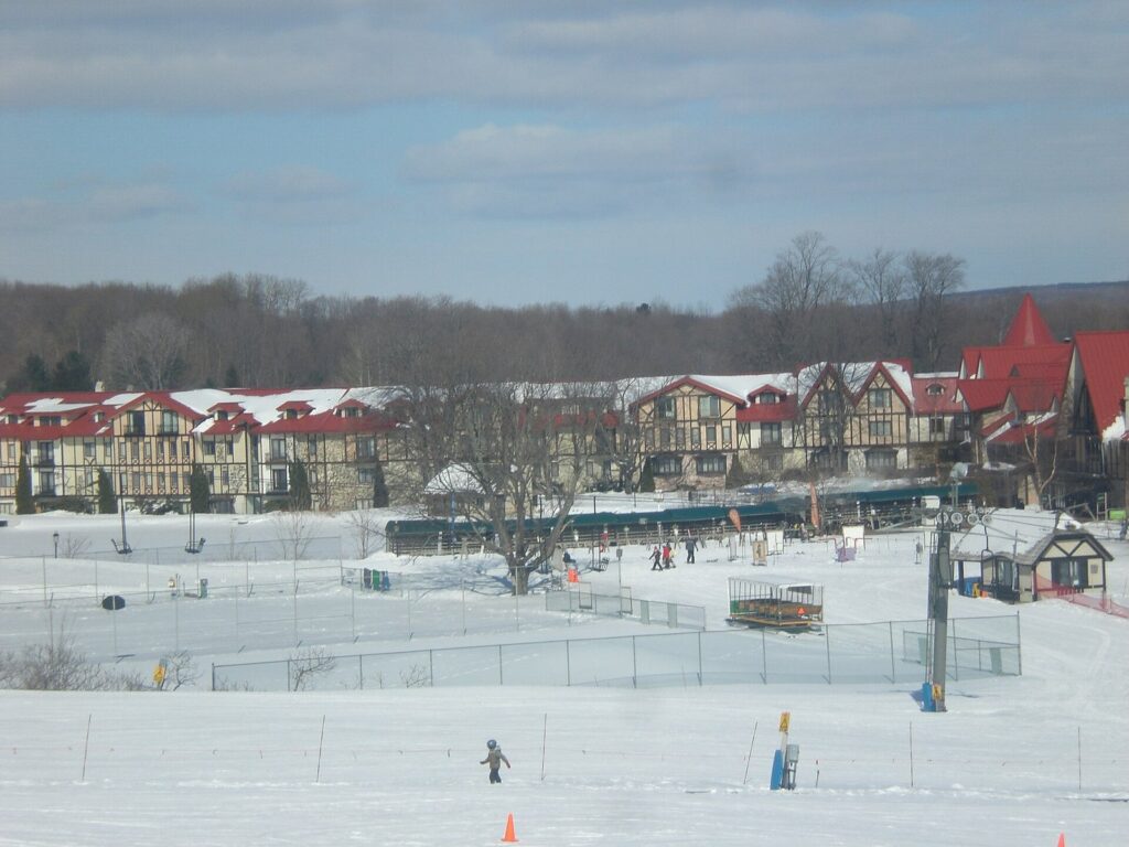 Snowy base area with a chairlift and ski school below a long, red-roofed timbered lodge