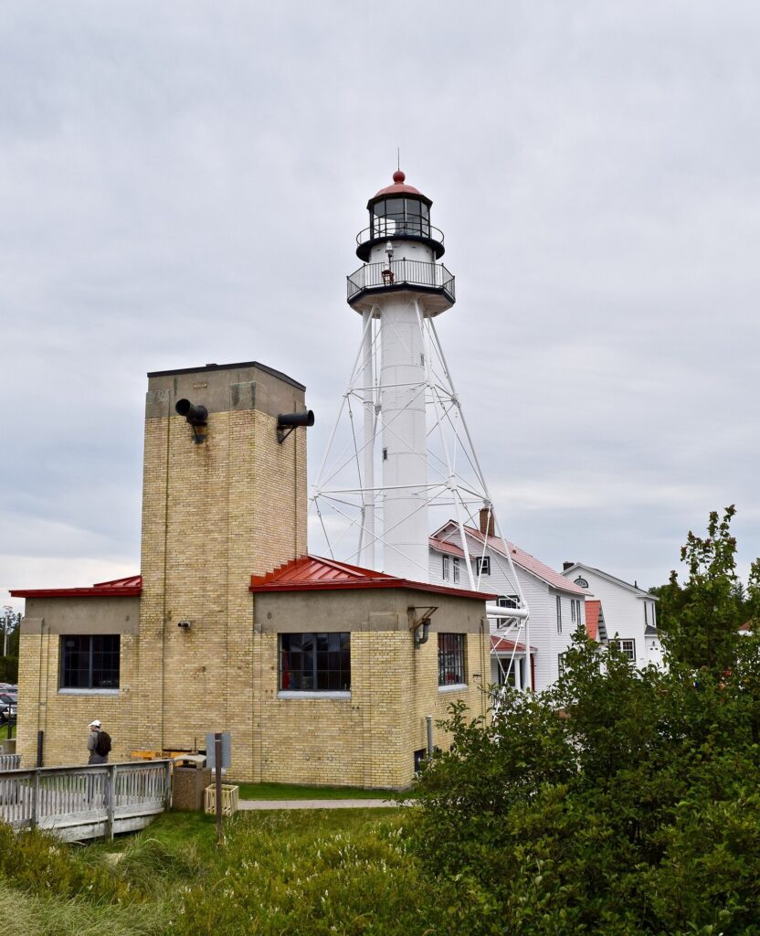 White steel-legged lighthouse tower rising behind a tan brick fog signal building at Whitefish Point