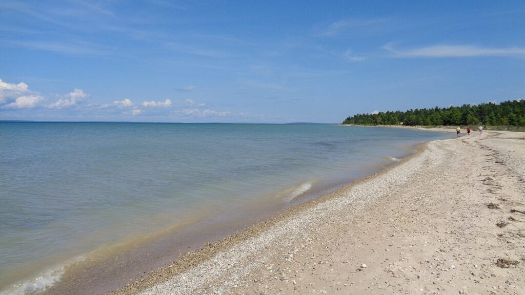 Sandy, pebbly Lake Michigan beach curving toward a wooded point under a blue sky with scattered clouds